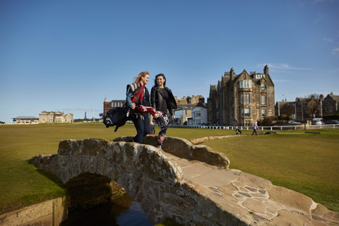 Two young women carrying golf clubs walking over the small Swilcan bridge at St Andrews golf course