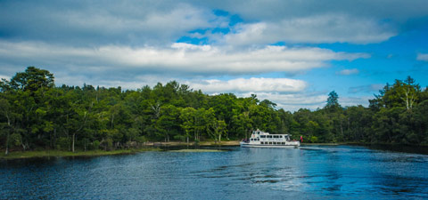 A white double-deck boat stops at a tree-lined bay while cruising Loch Lomond