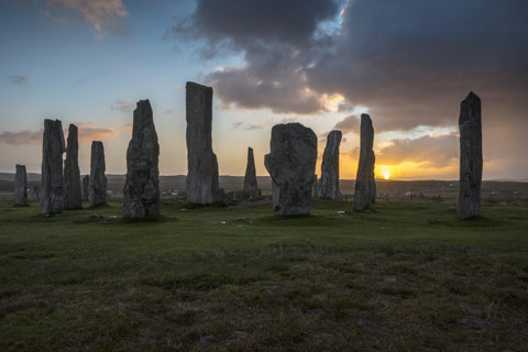 Callanish Standing Stones - group of grey stones in a field with the sun setting in clouds in background