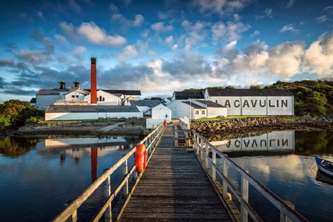 wooden bridge with railings leading over calm water  to low white buildings showing writing Lagavulin Distillery