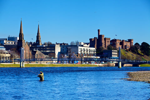 man fishing in deep blue river with Inverness Castle and other buildings in the background